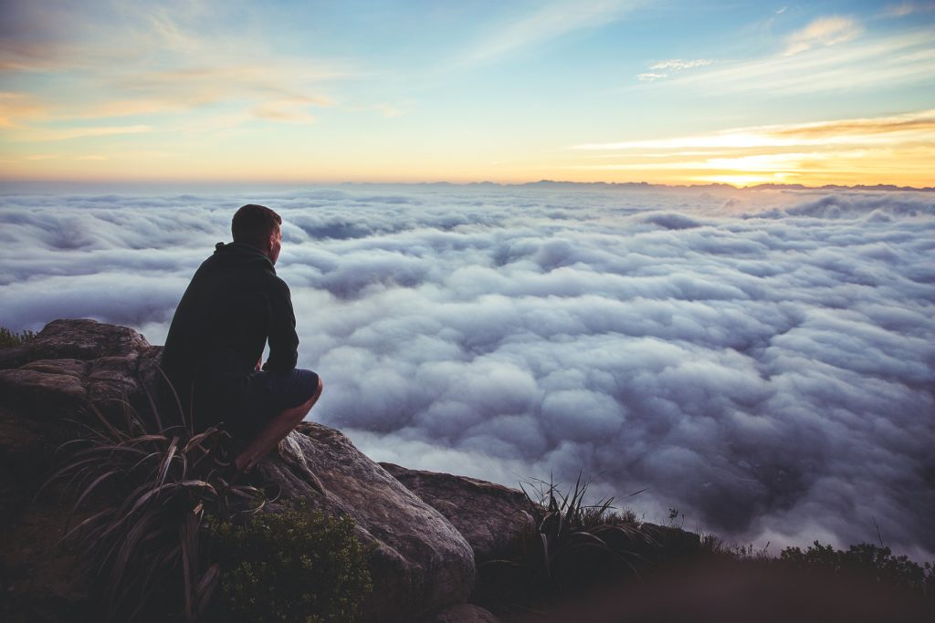 man on top of mountain looking at clouds below him